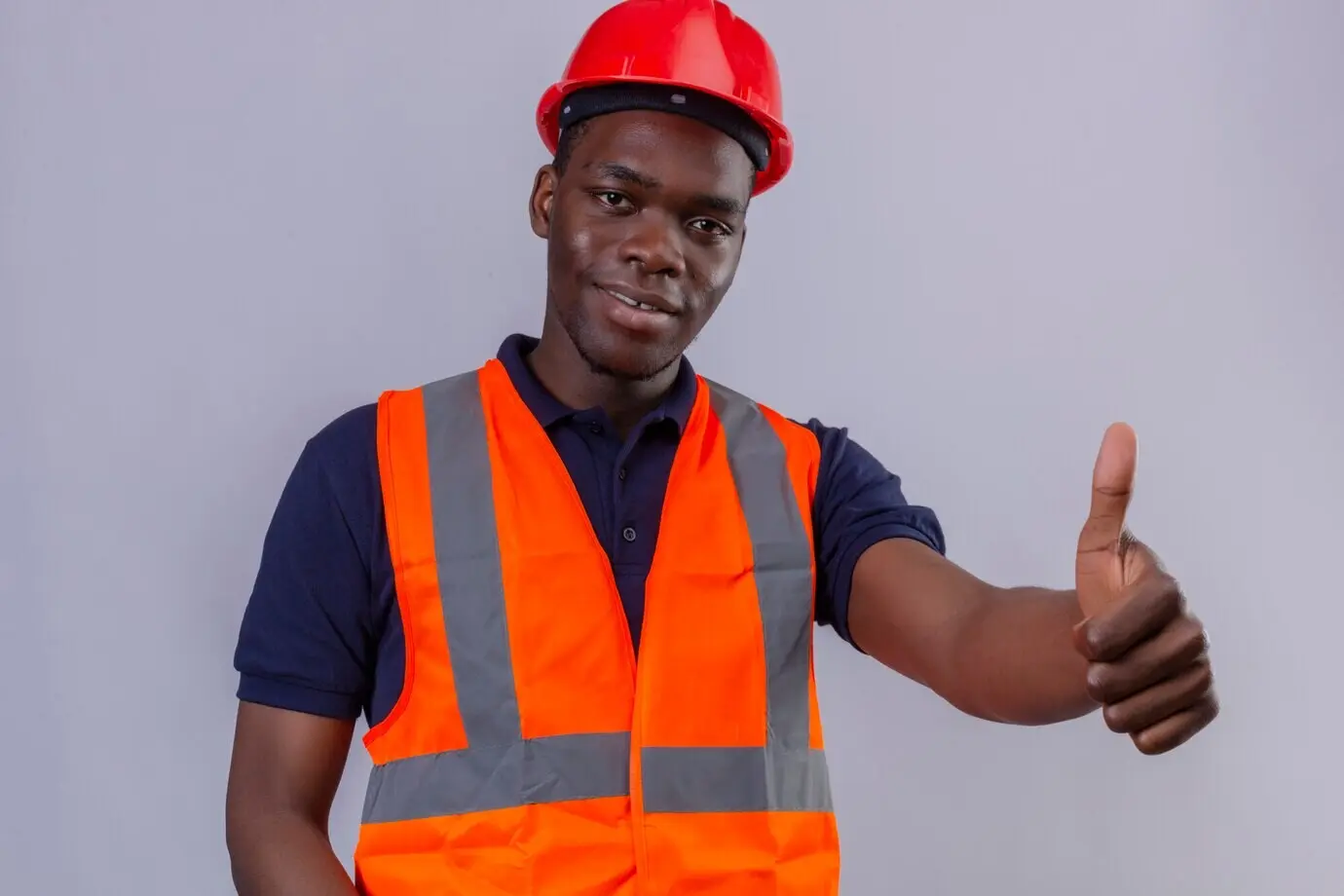 A young African American male builder wearing a construction vest and safety helmet, smiling and giving a thumbs-up while standing.