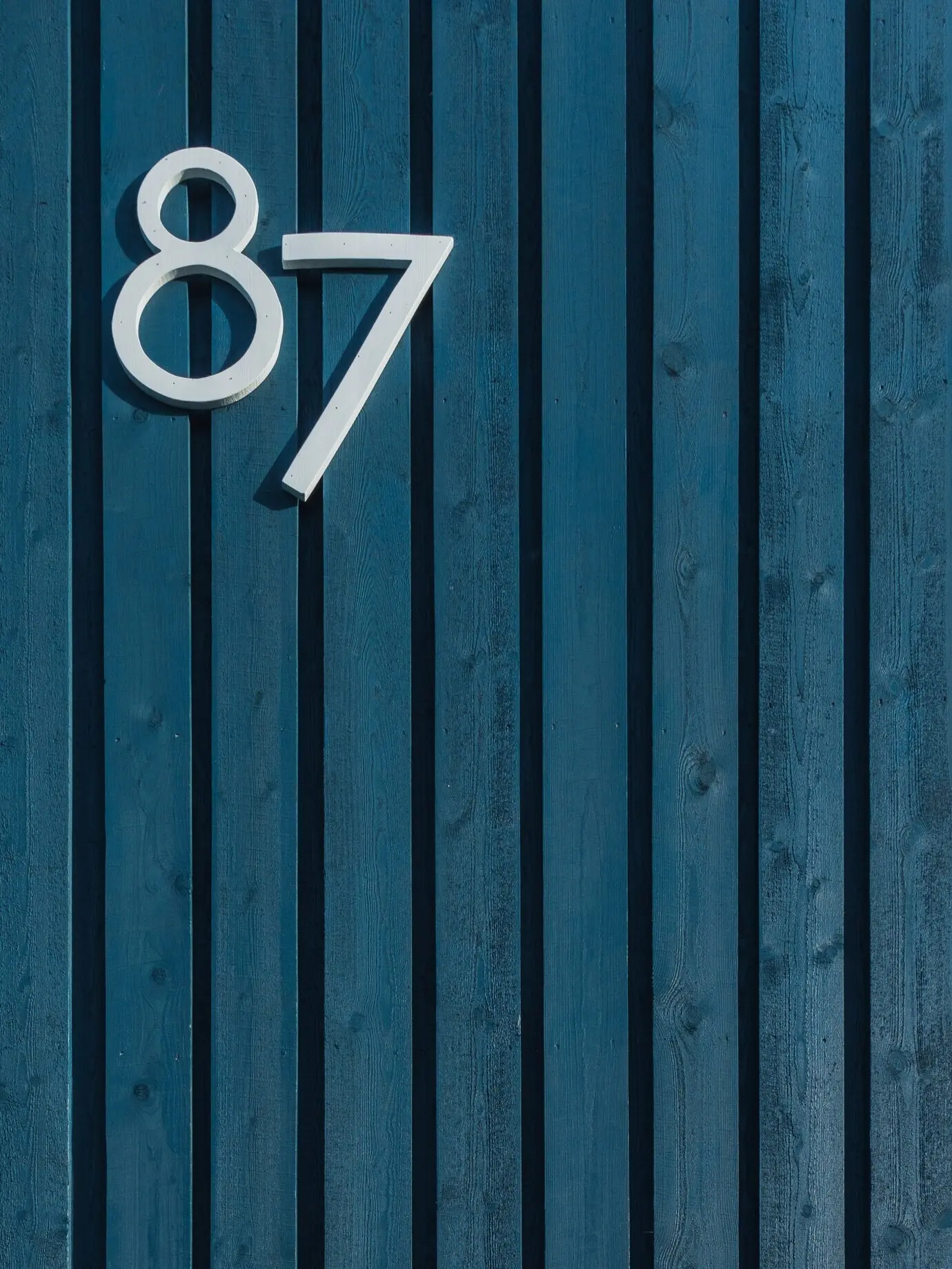 Vertical view of a blue wooden wall with vertically arranged sticks and the white number eighty-seven.