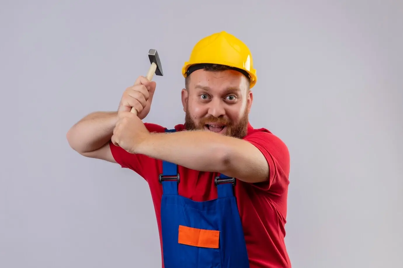 A young, bearded construction worker in uniform and a safety helmet swings a hammer, looking at the camera and smiling.